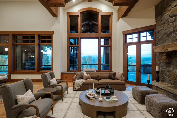 Vaulted ceiling featuring exposed timber beams, full-height windows with wood trim, floor-to-ceiling natural stone fireplace, and wood-finish flooring