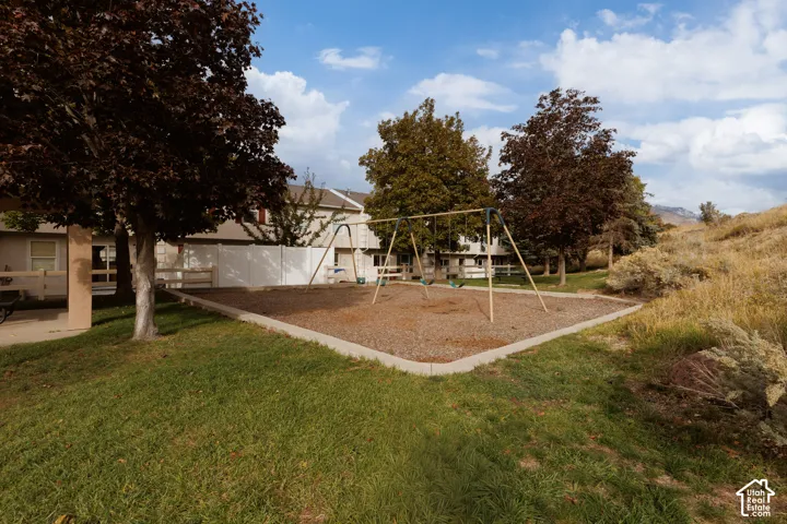 Outdoor common area featuring a swing set, wood-chip ground cover, manicured lawn, and mature trees with mountain backdrop