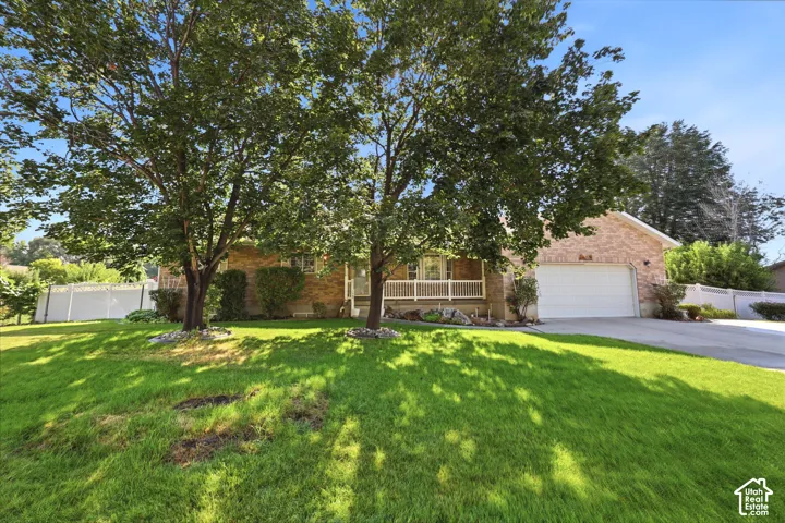 Brick exterior, covered front porch with white railing, attached two-car garage, mature shade trees, and perimeter vinyl fencing