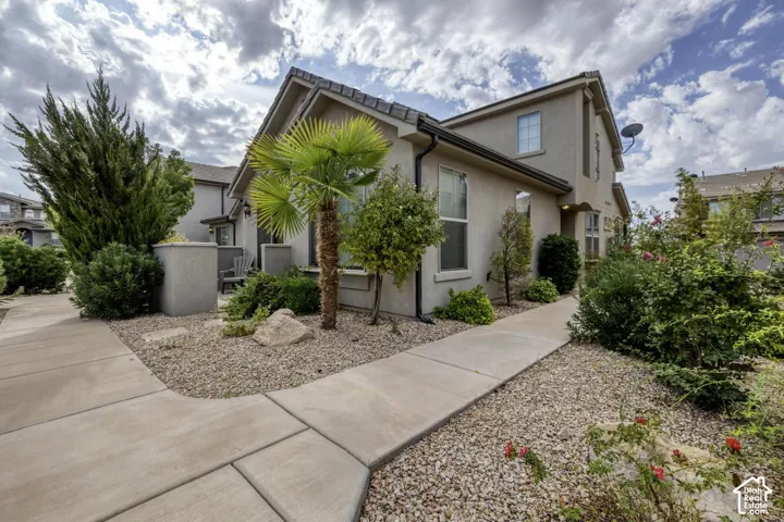 Stucco exterior featuring dual-pitched rooflines, concrete walkway, and landscaped desert-style gardens with native shrubbery and palm plantings