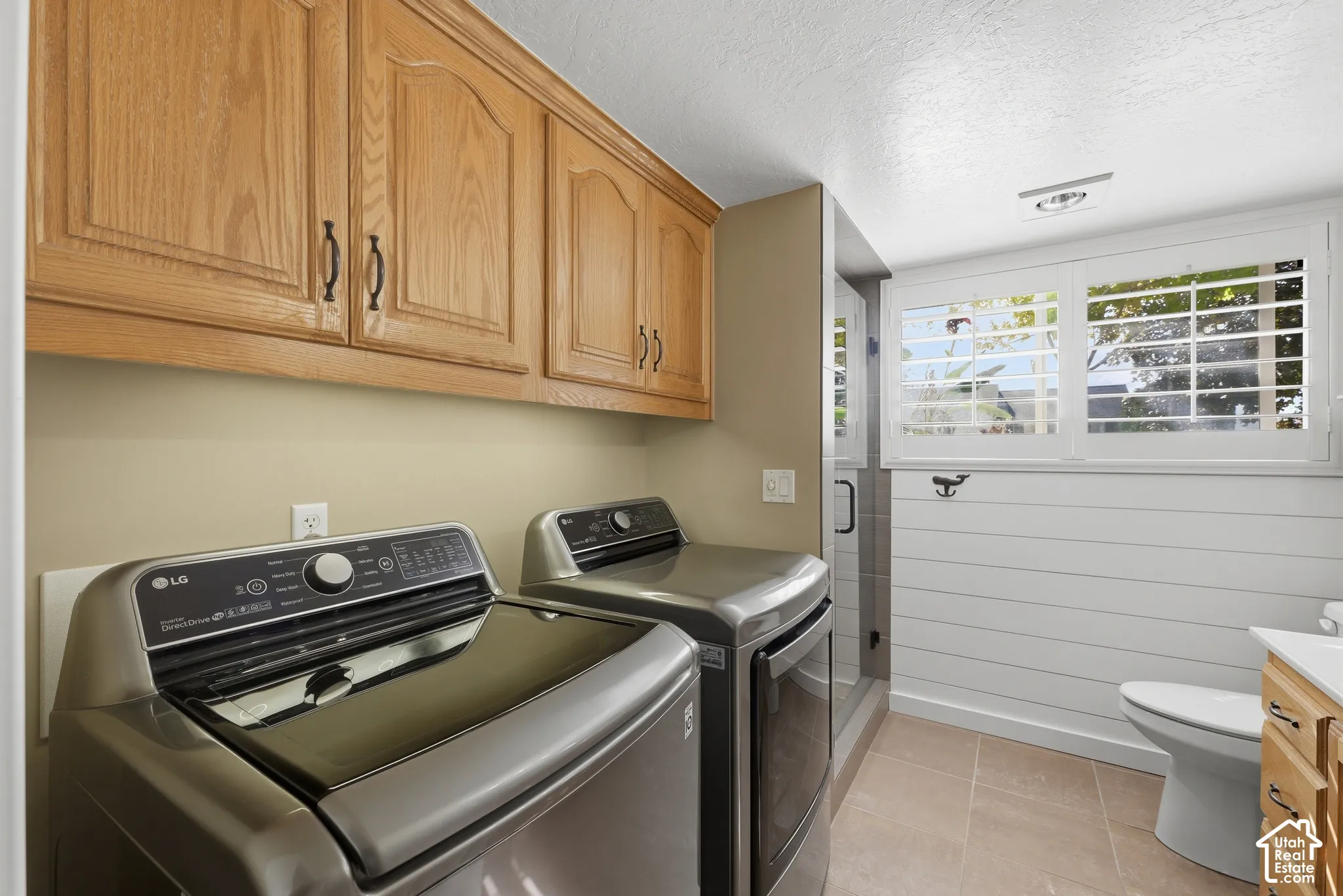 Laundry room featuring wood-finish upper cabinetry, white horizontal wall paneling, tiled flooring, and plantation shutters
