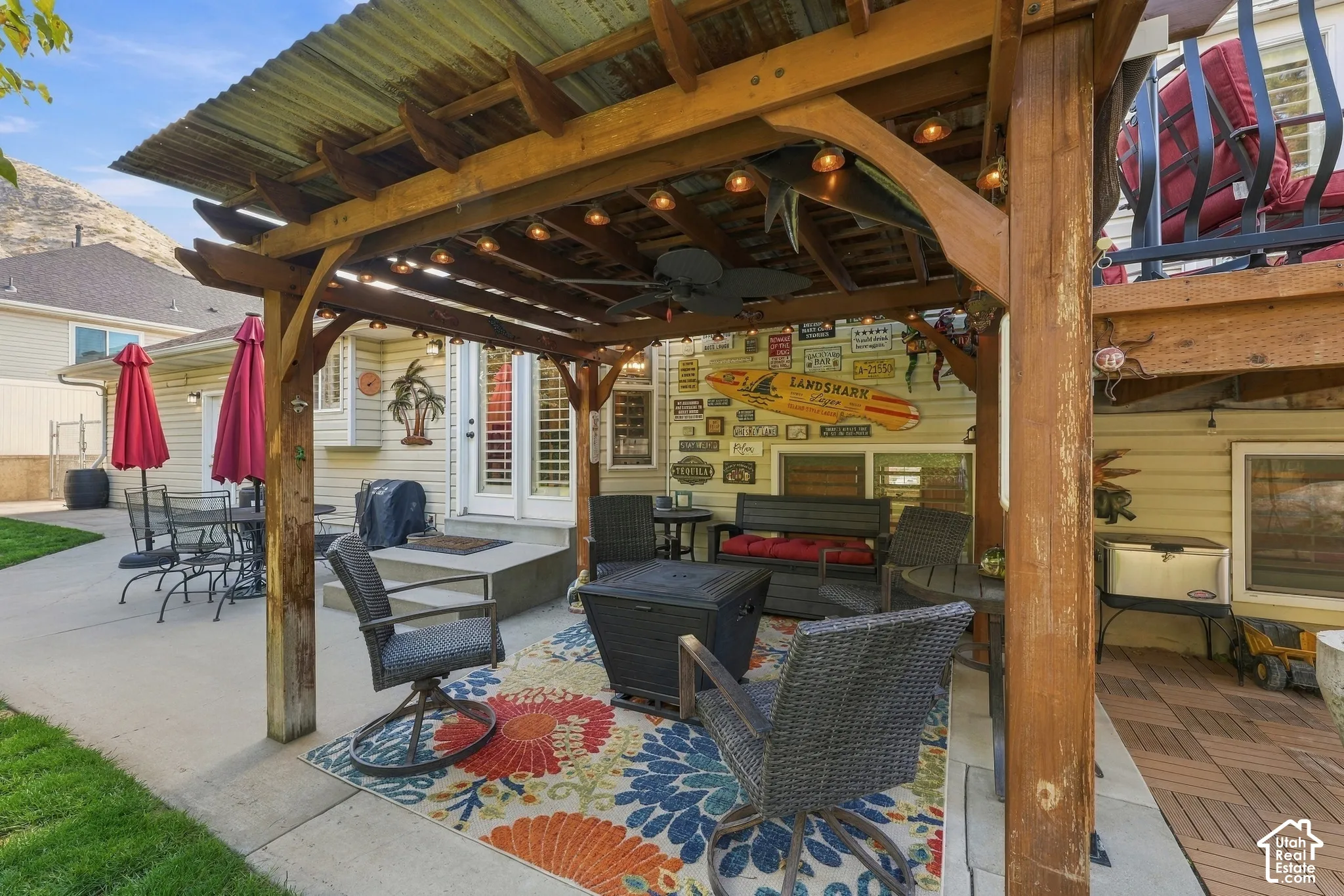 Covered outdoor pergola with corrugated metal roof, integrated ceiling fan, and string lighting, featuring a concrete patio surface and multi-level composite decking