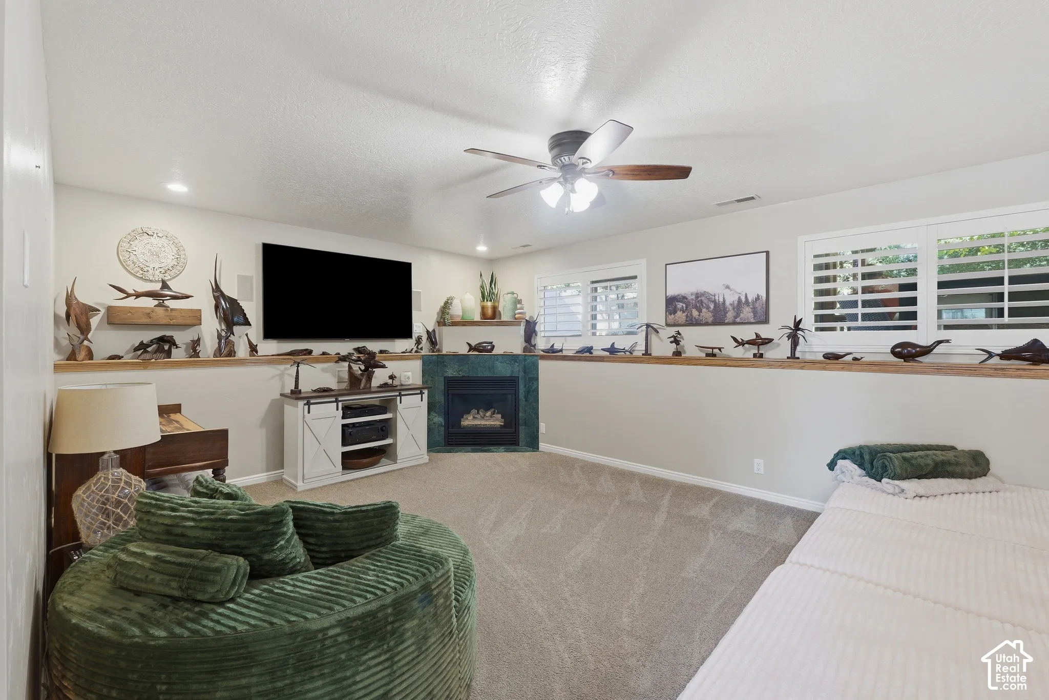 Corner fireplace with a tiled surround, carpeted flooring, built-in wooden shelving ledge, ceiling fan with light fixture, and plantation shutters
