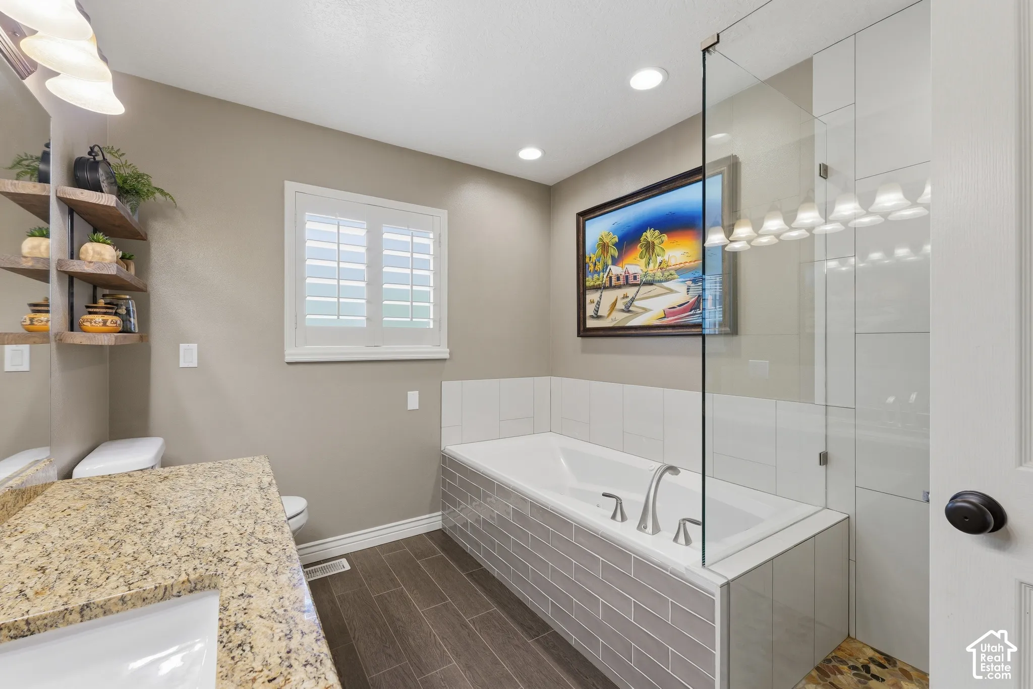 Bathroom featuring wood-finish flooring, a soaking tub with integrated tile surround, glass shower panel, stone-finish vanity countertop, and wooden wall-mounted shelving