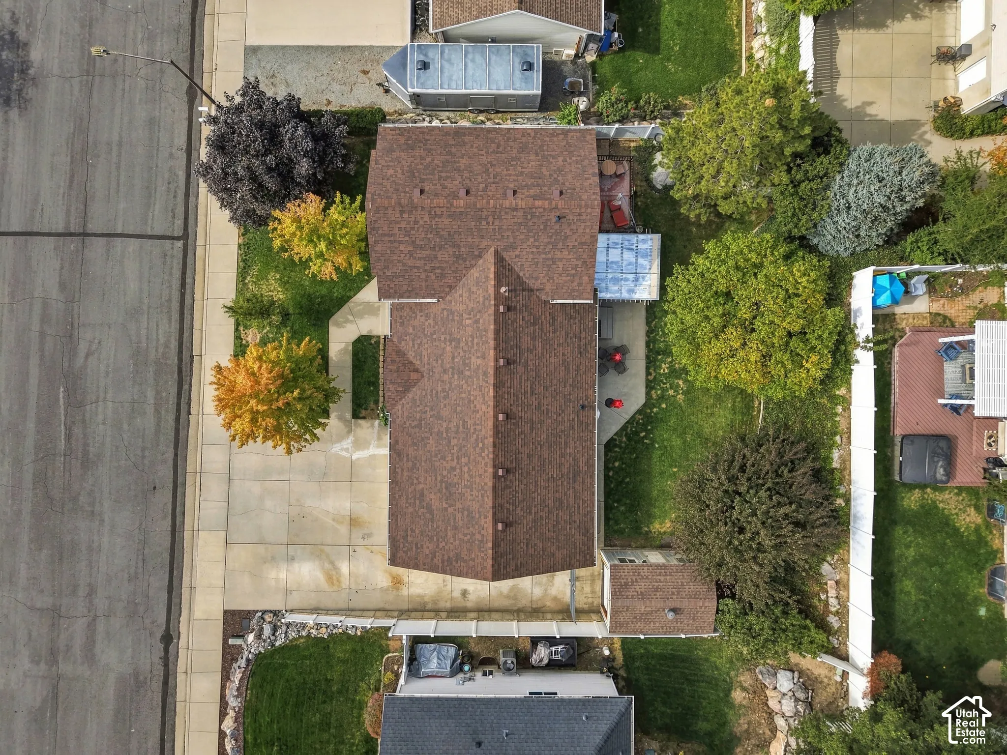 Aerial perspective of a property featuring a concrete driveway, multi-gabled shingle roof, established trees, and fenced rear lawn