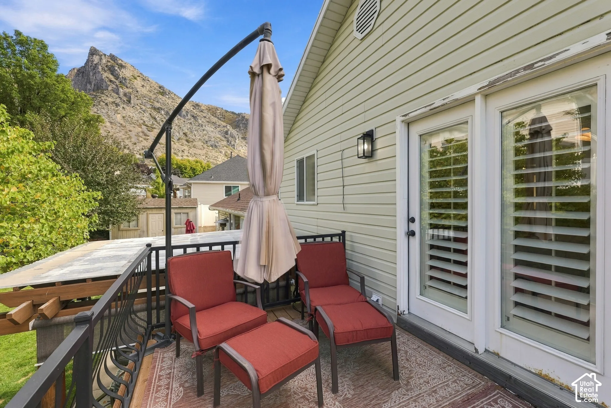Elevated deck featuring black metal railing, horizontal siding, exterior wall-mounted lighting, and glass-paneled doors with internal plantation shutters, set against a mountainous backdrop