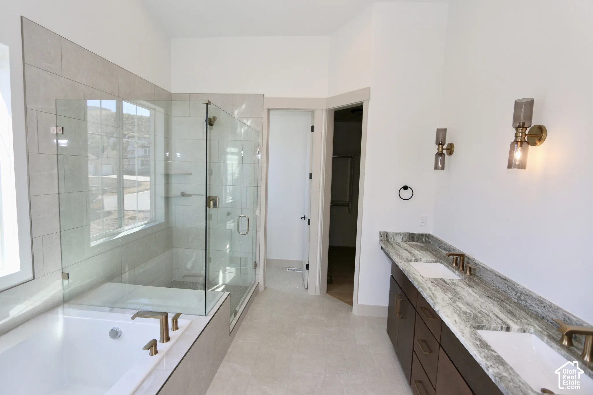 Master bathroom featuring a double-sink vanity with natural stone countertops, dark-finish cabinetry, a glass-enclosed walk-in shower, a soaking tub, and wall-mounted light fixtures