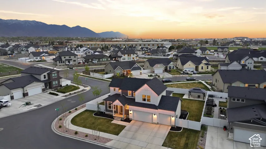 Aerial perspective of a two-story residence featuring a dual-car garage, white siding with dark trim accents, a dark asphalt shingle roof, a fenced backyard with a gazebo, and mountainous scenery