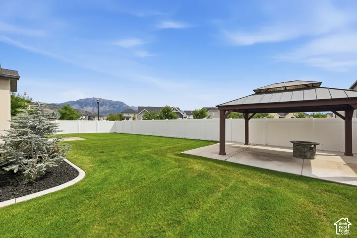Expansive backyard featuring a manicured lawn, white vinyl fencing, concrete patio, metal-roof gazebo, and mountain backdrop