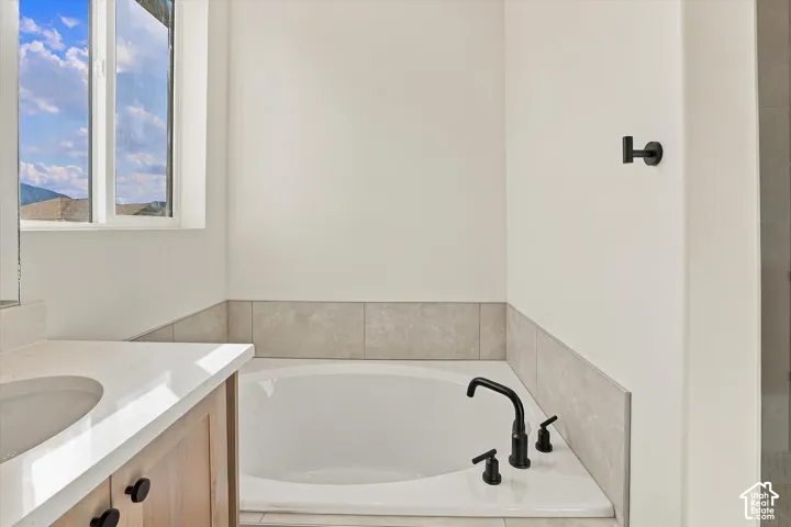 Built-in soaking tub with matte black deck-mounted faucet, stone-surround tile, light wood-finish cabinetry, quartz-style countertop, and matte black wall-mounted hardware