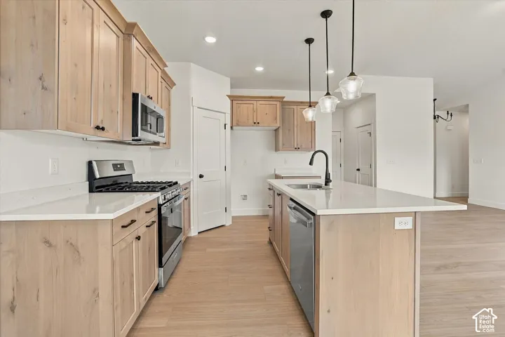 Kitchen featuring light wood-finish cabinetry, quartz countertops, stainless steel appliances, a central island with a matte black faucet, and pendant lighting