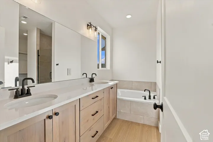Dual-sink vanity with light-tone wood cabinetry, solid-surface countertops, matte black plumbing fixtures, and a tiled soaking tub surround
