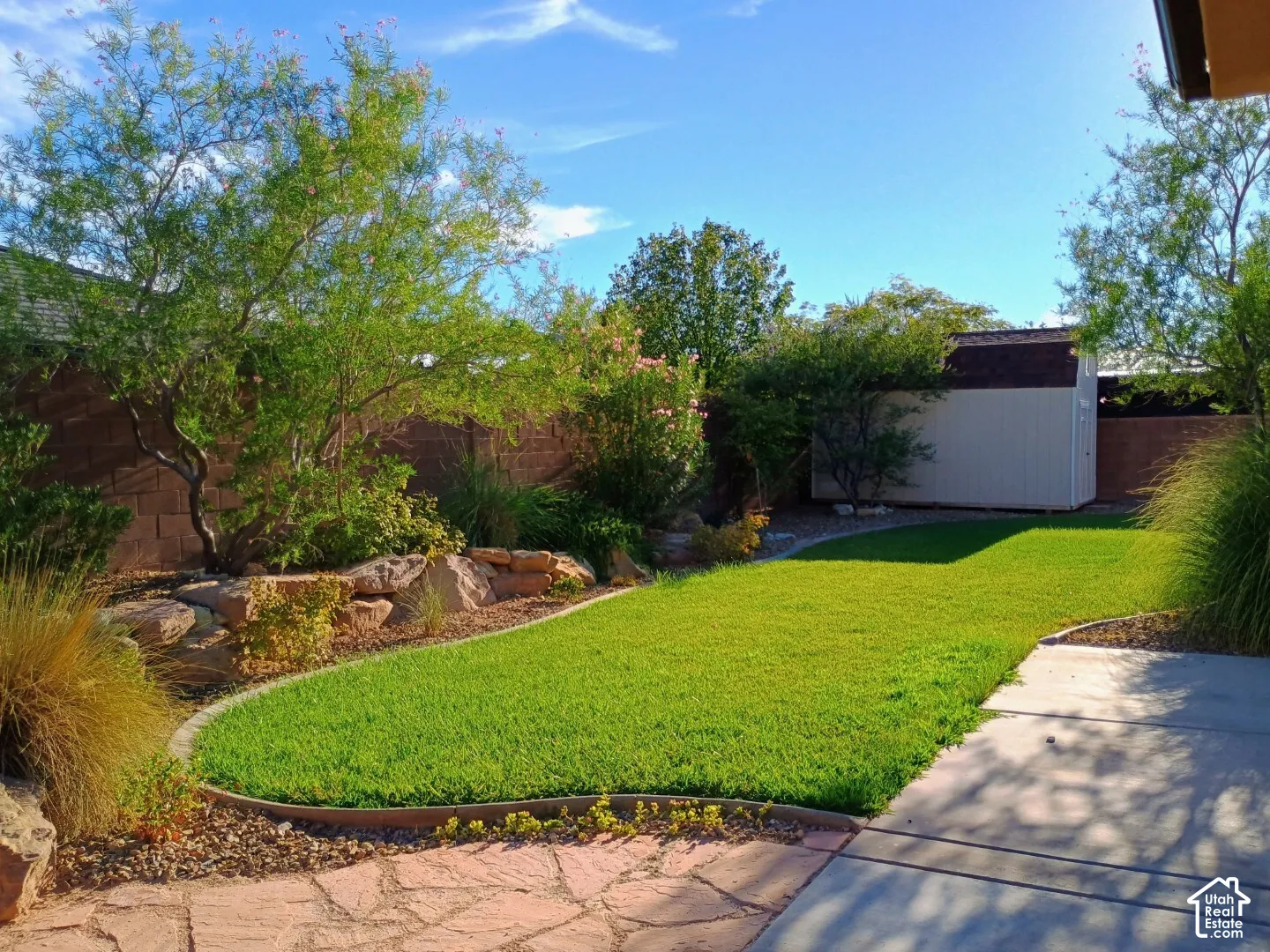 Landscaped backyard featuring a natural stone patio, manicured lawn, tiered stone retaining wall, mature trees, and an exterior storage structure