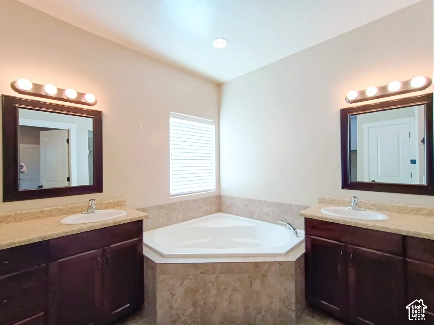 Dual vanities featuring dark wood-finish cabinetry, stone-texture countertops, undermount oval sinks, and framed mirrors, paired with a corner soaking tub encased in decorative tile