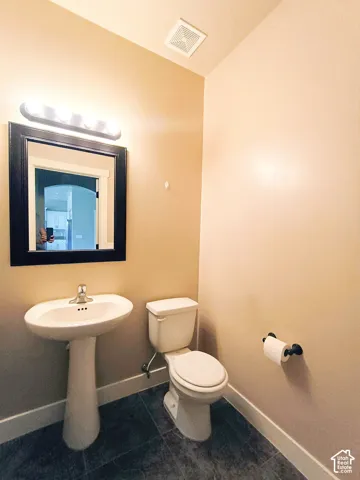 Pedestal sink with chrome faucet, framed mirror, wall-mounted light fixture, and dark-toned tile flooring