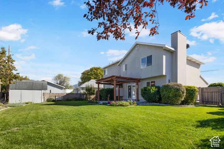 Exterior rear elevation featuring a stucco facade, a prominent chimney, an attached wood-frame pergola, a large fenced backyard with a lawn, and a detached storage shed