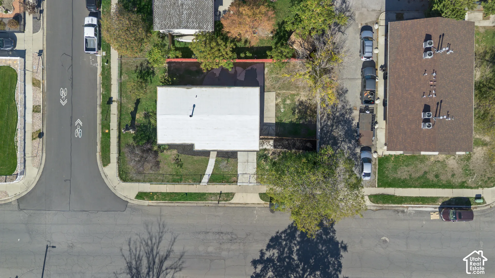 Aerial perspective of a corner lot property featuring a light-toned roof, concrete walkway, detached pavement parking area, and mature trees