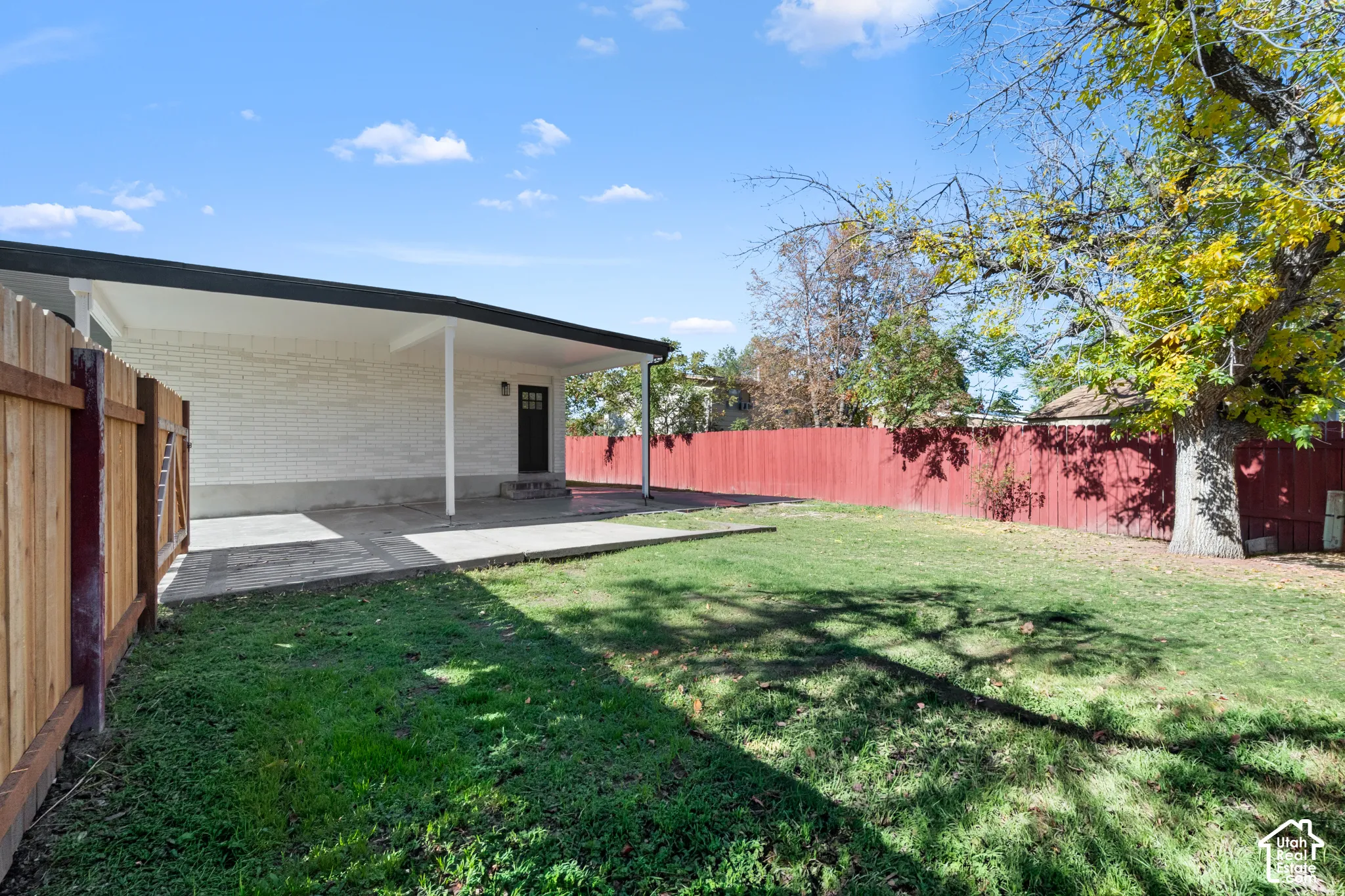 Covered concrete patio, white brick exterior, mature trees, and wooden perimeter fencing
