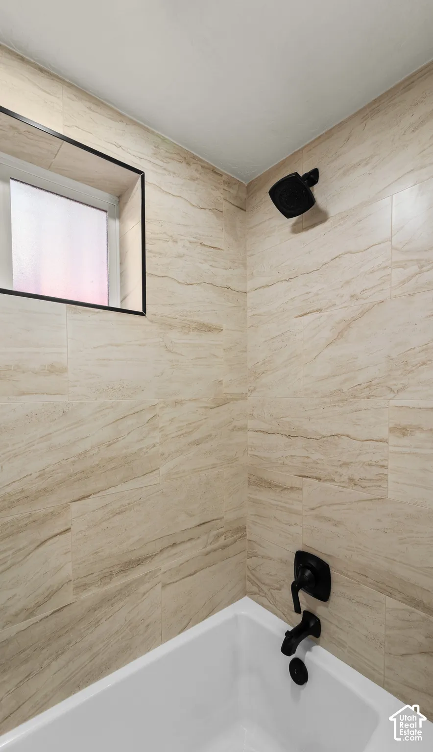Full-height beige tile surround with natural veining, matte black showerhead, matching matte black plumbing fixtures, and a recessed window with a black frame