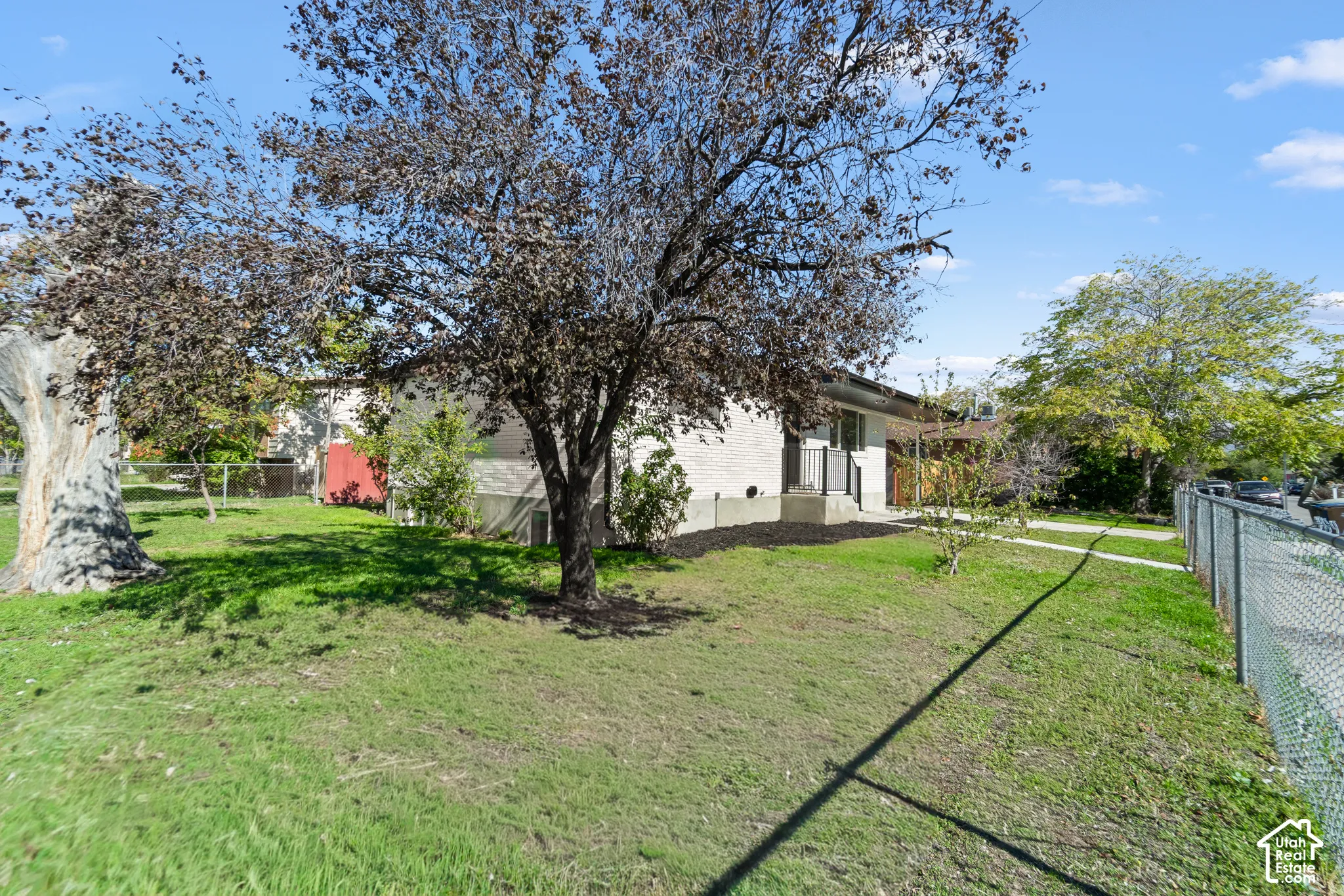 Exterior view featuring light-colored brick siding, mature trees, and a chain-link perimeter fence