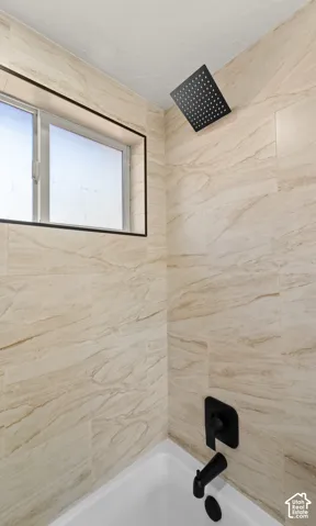 Shower enclosure featuring large-format beige tile surround, square matte black rainfall showerhead, matte black wall-mounted faucet, and window with dark trim