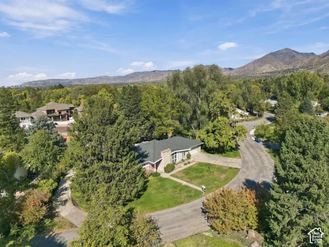 Single-story residence featuring a shingle roof, masonry chimney, concrete driveway, and established landscape situated against a mountain backdrop