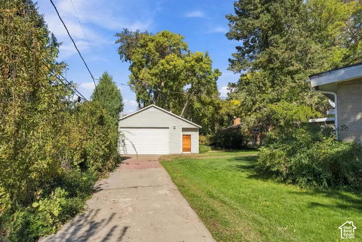Detached garage featuring light-colored horizontal siding, a single-bay overhead door, and a dedicated side-entry door, complemented by a paved driveway and mature trees