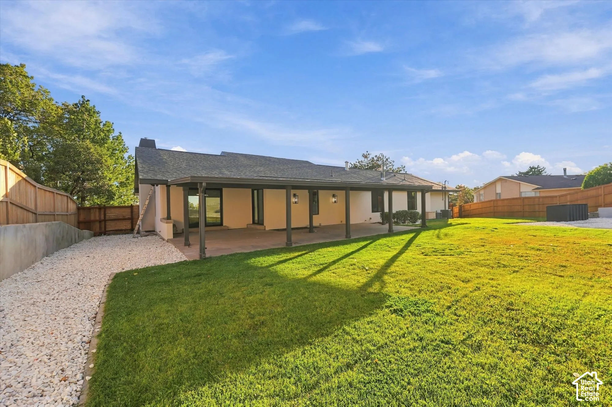 Rear exterior featuring a covered patio, expansive lawn, stone-surfaced side yard, and perimeter wood fencing