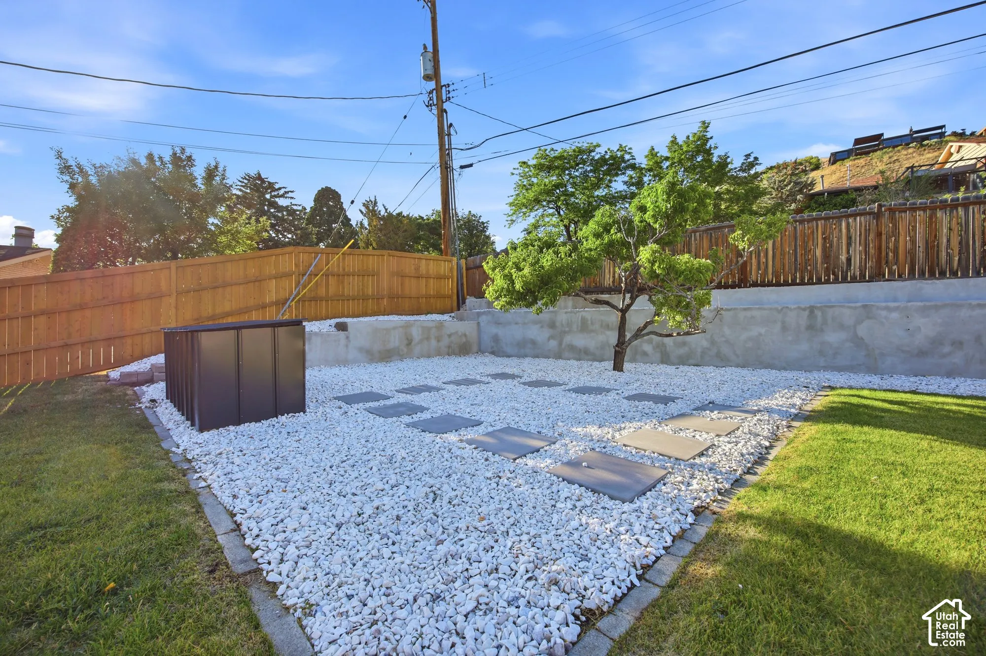 Backyard featuring white gravel landscaping, concrete retaining walls, integrated square stone pavers, wood fencing, and an established tree