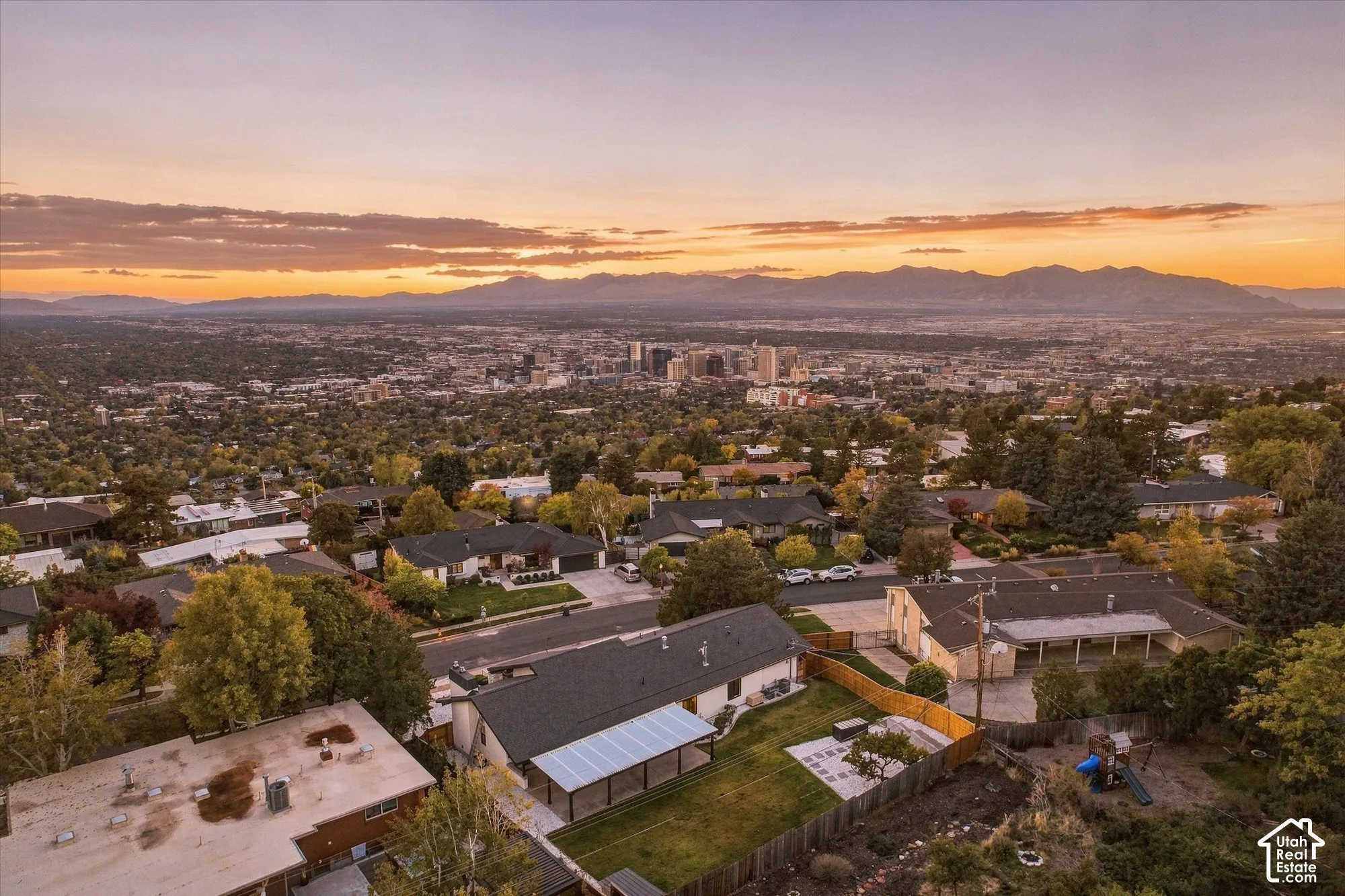 Aerial perspective featuring a shingled roof, an expansive rear patio with a covered pergola structure, a paved hardscape area, and a prominent mountain range backdrop