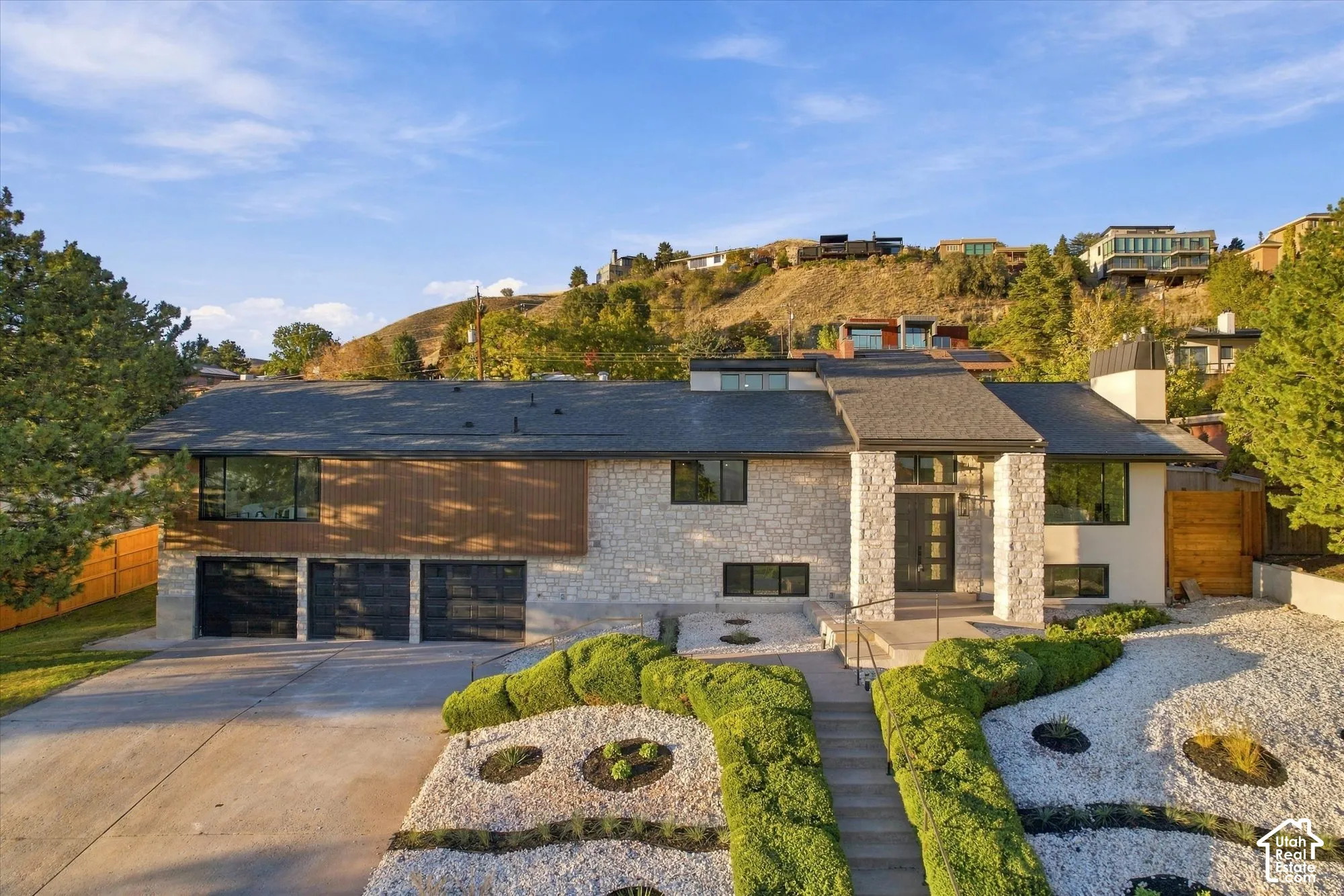 Multi-level exterior featuring stone veneer siding, horizontal wood-slat accents, a three-car garage, and tiered landscaping