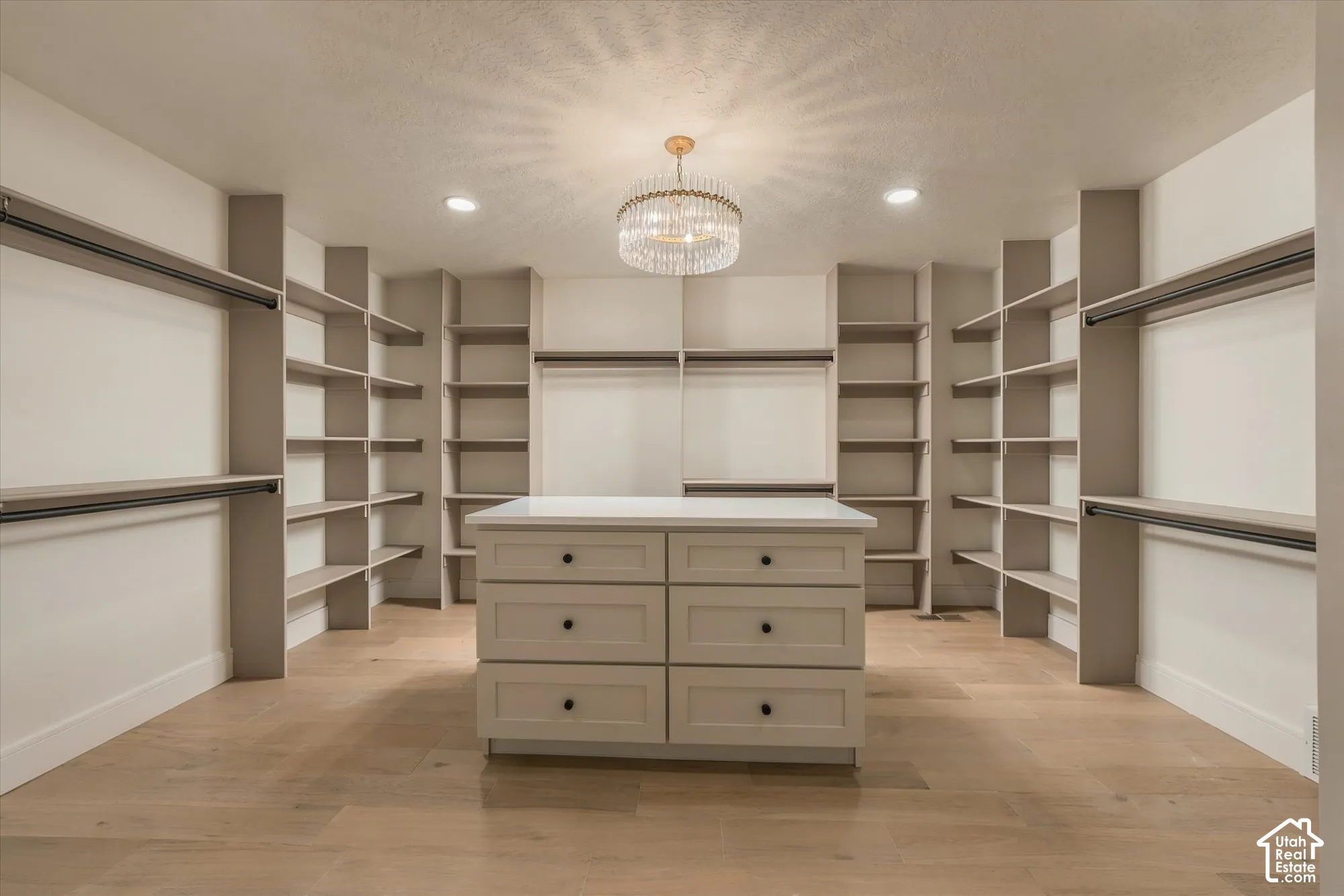 Walk-in closet featuring floor-to-ceiling built-in shelving, wood-finish flooring, a central island with six drawers and a solid surface countertop, and a tiered crystal chandelier