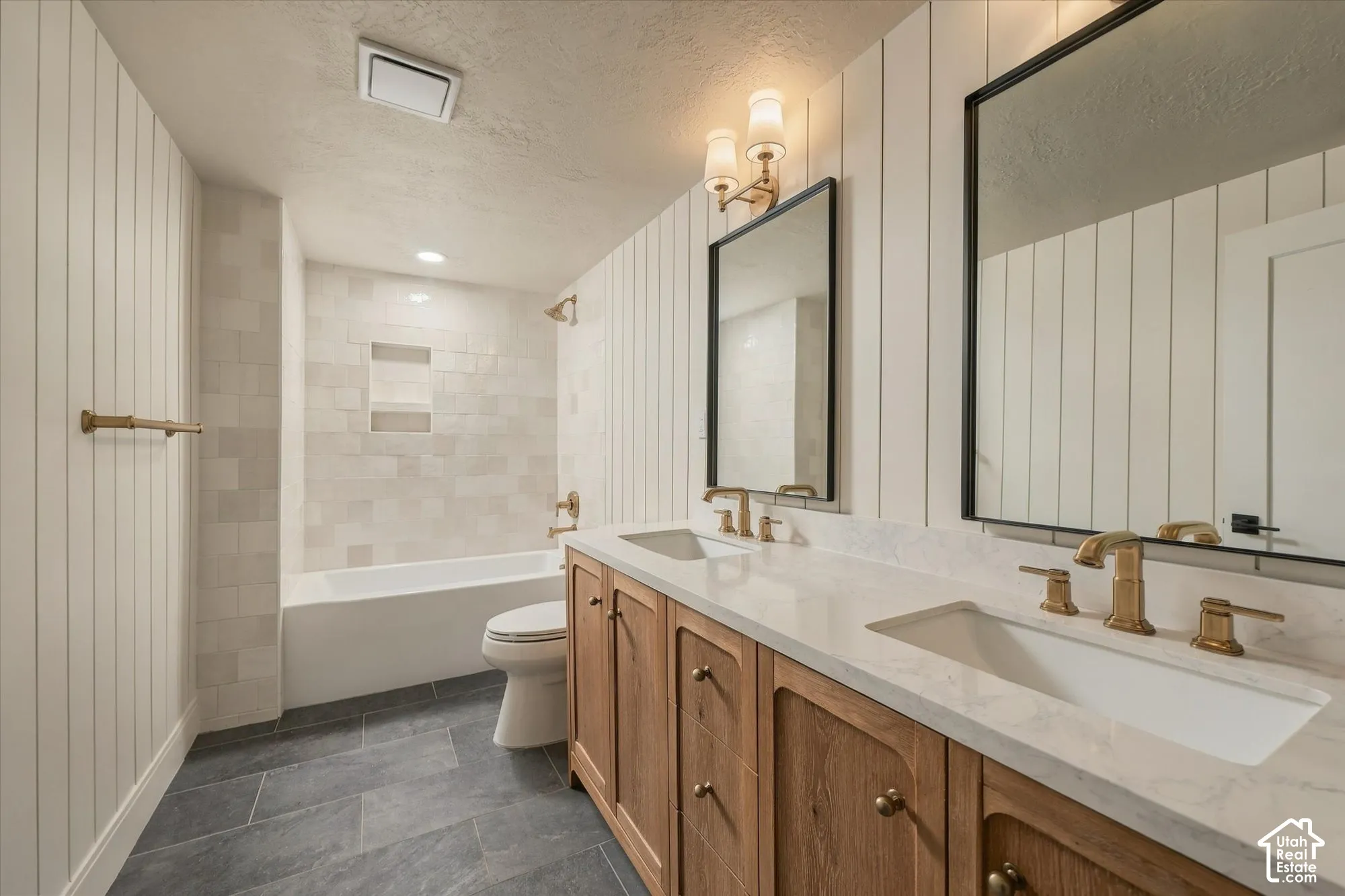 Double-sink vanity with light-tone wood-finish cabinetry, stone-surface countertop, vertical shiplap wall paneling, and matte-gold finish fixtures