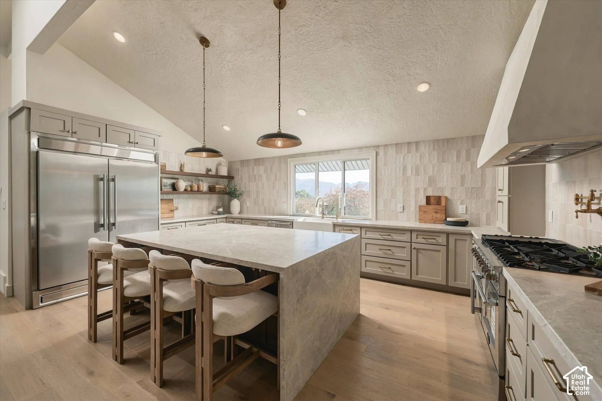 Kitchen featuring a waterfall-edge center island, stone-surfaced countertops, custom cabinetry with brushed hardware, stainless steel professional-grade appliances, and wood-finish flooring