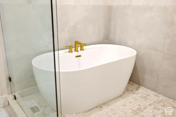 Freestanding soaking tub with gold-tone faucet, complemented by large-format neutral wall tile, decorative geometric floor tile, and a glass-enclosed shower