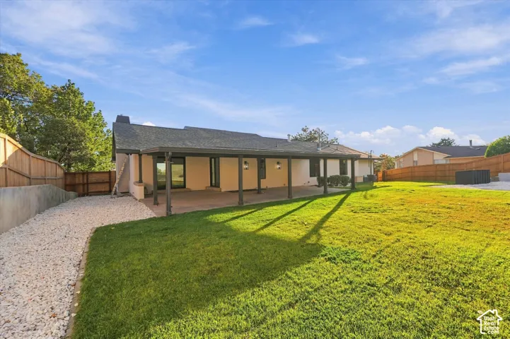 Rear exterior featuring a covered patio, expansive lawn, stone-surfaced side yard, and perimeter wood fencing
