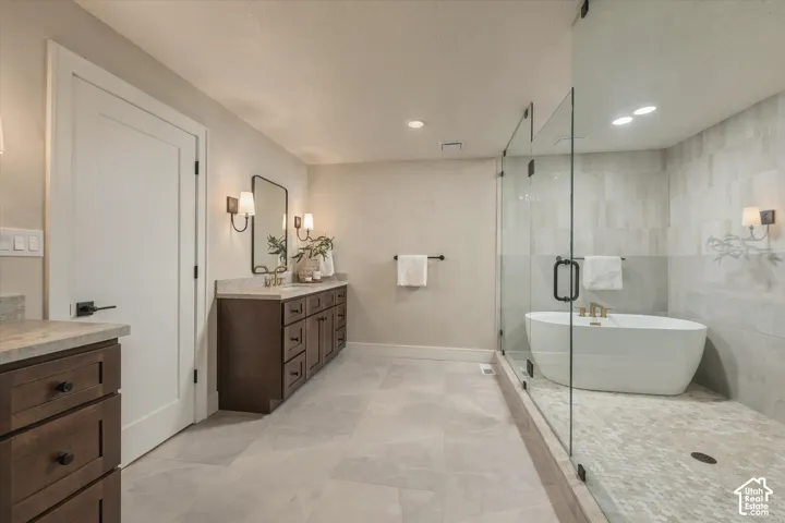 Dark wood cabinetry with stone-finish countertops, glass-enclosed wet room, freestanding soaking tub, large-format neutral floor tile, and dual wall-mounted light fixtures