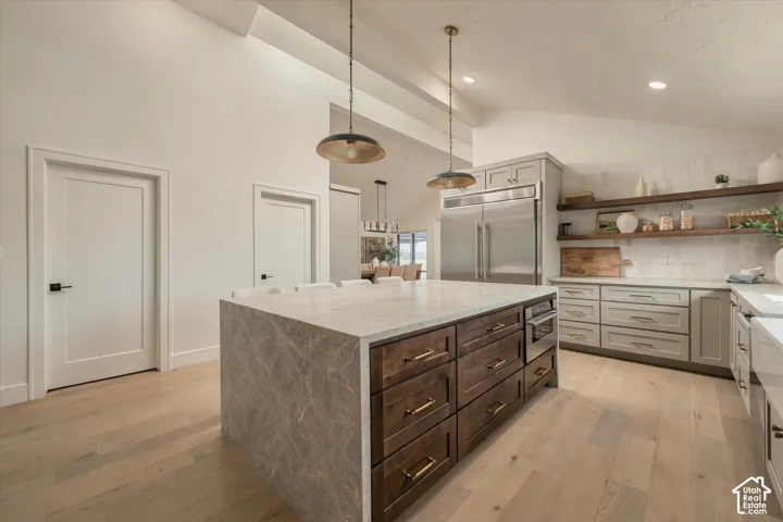 Kitchen featuring a central island with stone-finish waterfall countertop, wood-finish flooring, stainless steel built-in refrigerator, custom cabinetry with metallic hardware, and open wood shelving