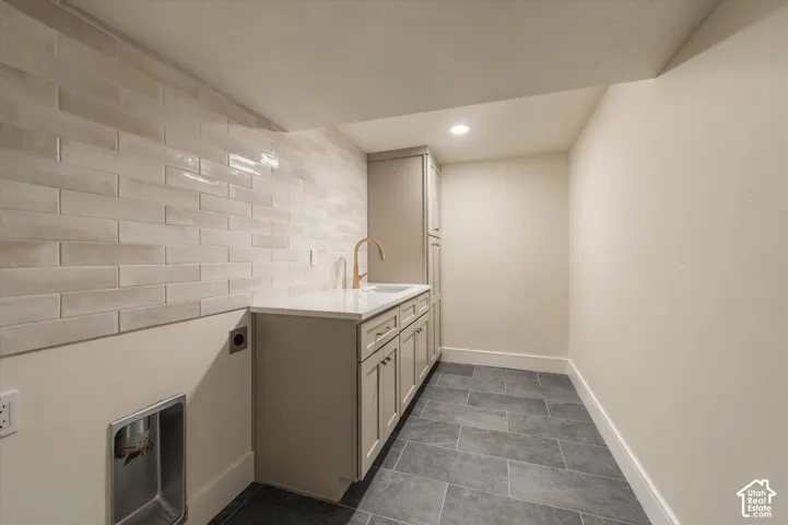Laundry space featuring gray stone-finish flooring, custom shaker-style cabinetry, a solid-surface countertop, a neutral subway tile backsplash, and a tall arched-neck faucet