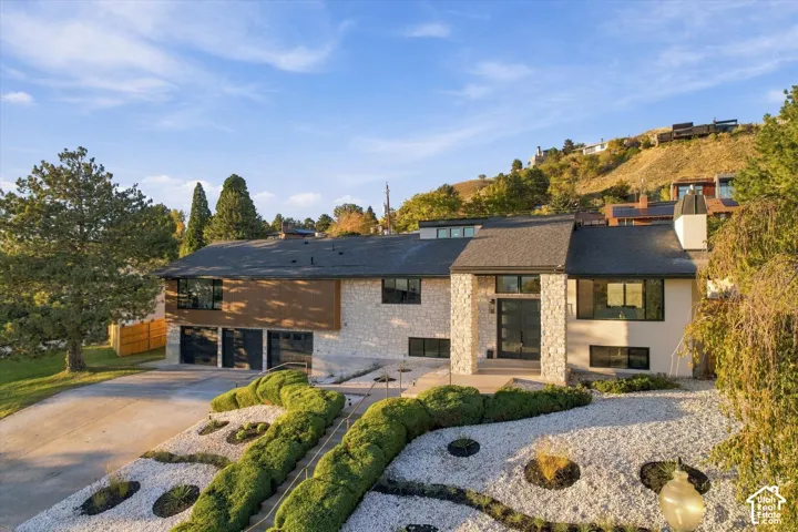 Contemporary split-level exterior featuring light stone masonry, dark wood-tone siding, a three-car garage, and tiered rock landscaping