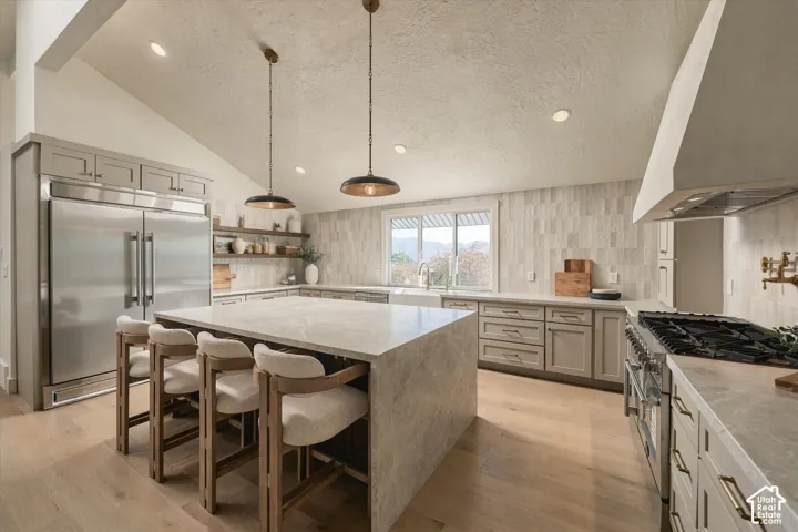 Kitchen featuring a waterfall-edge center island, stone-surfaced countertops, custom cabinetry with brushed hardware, stainless steel professional-grade appliances, and wood-finish flooring
