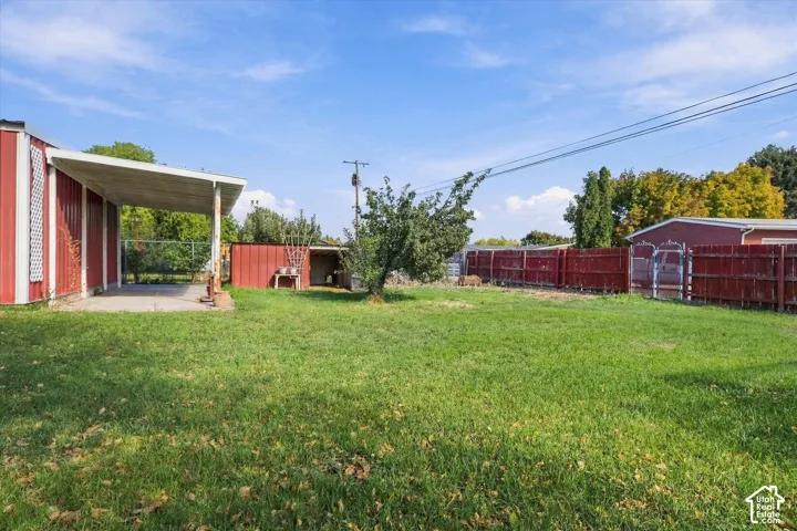 Large backyard featuring a concrete patio, metal-siding exterior structure, perimeter fencing, and mature trees