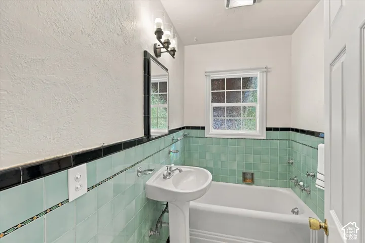 Bathroom featuring a porcelain pedestal sink, white paneled tub, and light green square tile wainscoting with a black decorative border