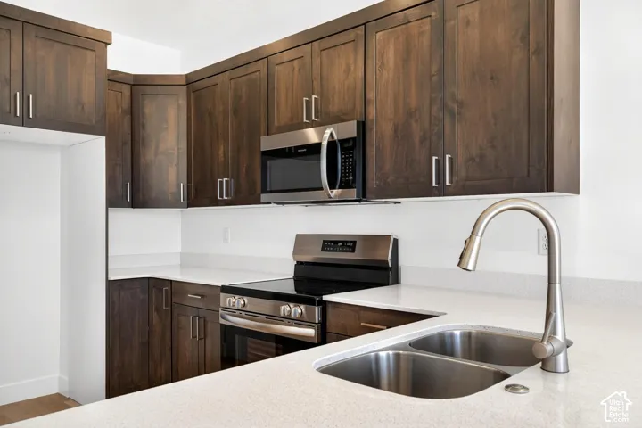 Kitchen featuring dark-toned wooden cabinetry, light-colored stone-finish countertops, stainless steel appliances, and a double-basin undermount sink with a gooseneck faucet