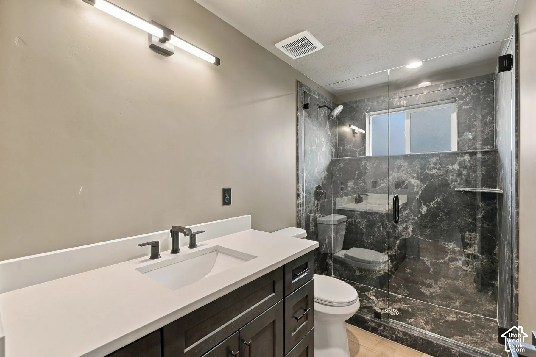 Bathroom featuring a dark wood-finish vanity with a light-toned solid surface countertop, matte black widespread faucet, linear wall-mounted lighting, and a walk-in shower with floor-to-ceiling stone-finish surround and frameless glass enclosure