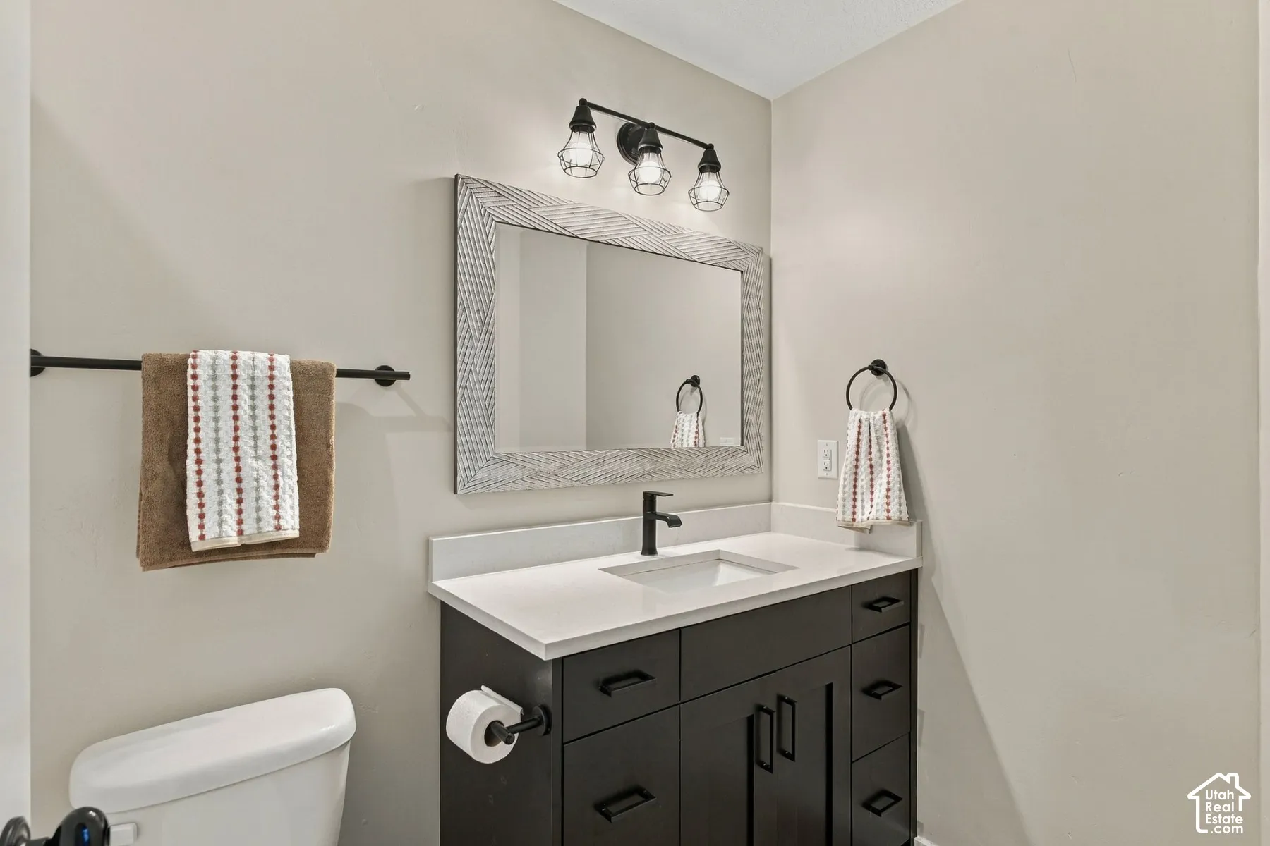 Dark-finish vanity featuring a solid surface countertop, an undermount sink, matte black hardware, a three-light fixture, and a framed mirror