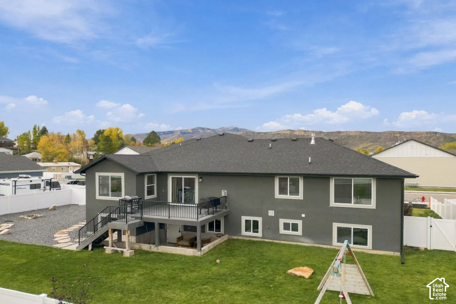Exterior featuring a gray stucco facade, elevated deck with metal railings, gravel and grass landscaping, white vinyl perimeter fencing, and a mountain backdrop