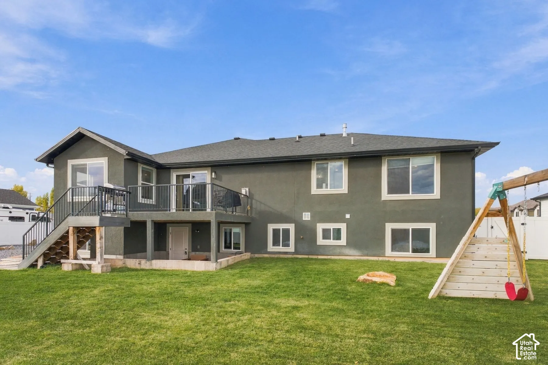 Rear exterior featuring a stucco facade, elevated deck with metal railing, staircase, and expansive grass lawn