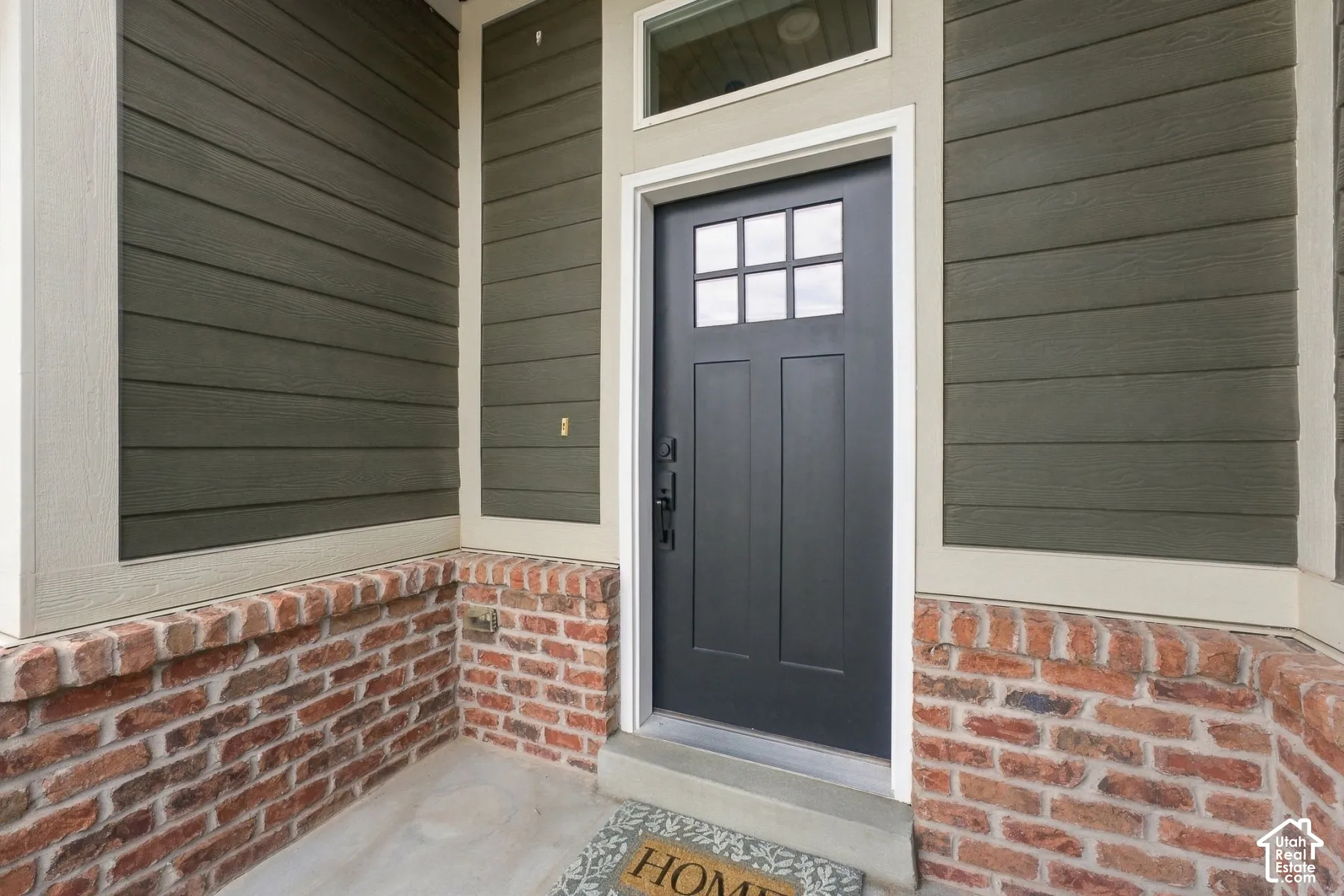 Entryway featuring a dark-toned door with a six-pane window insert, horizontal lap siding, a red brick base, and a transom window