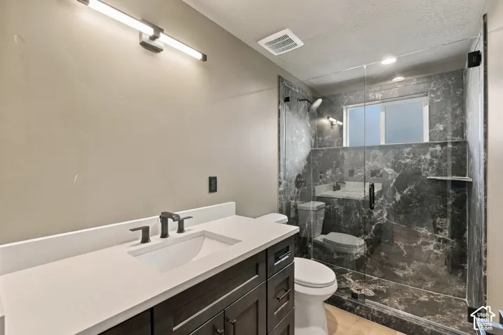 Bathroom featuring a dark wood-finish vanity with a light-toned solid surface countertop, matte black widespread faucet, linear wall-mounted lighting, and a walk-in shower with floor-to-ceiling stone-finish surround and frameless glass enclosure